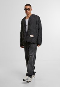 Black quilted jacket with a relaxed fit, featuring a button closure, one pocket, and a branded label on the hem. Worn with gray pants and sneakers.