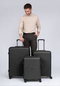 Trois valises rigides noires de tailles variées. Surface texturée avec des lignes diagonales et des poignées rétractables. Roues lisses pour un transport facile.