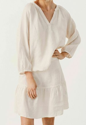 Vrouw draagt een losse, lange mouwen, crèmekleurige blouse en bijpassende knielange rok, staand met één hand op de heup.