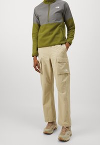 Giacca in pile con zip a quattro quarti in verde e grigio; pantaloni cargo beige con due grandi tasche laterali; scarpe da trekking beige con accenti arancioni.
