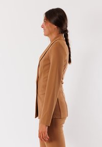 Blazer marrone aderente realizzato in tessuto texturizzato, con design a un bottone, rever a lancia e fessure sui lati. Abbinato a pantaloni coordinati.