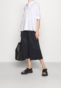 Camicetta bianca con maniche corte, abbinata a pantaloni neri a gamba larga e cropped, e sandali neri senza chiusura. Borsa nera con manico.