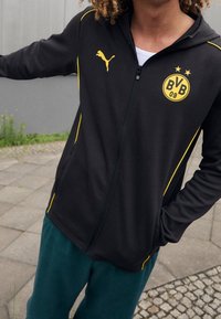 Persona che indossa una giacca nera con zip e logo giallo Puma e emblem BVB 09, in piedi su un terreno pavimentato con un braccio esteso.