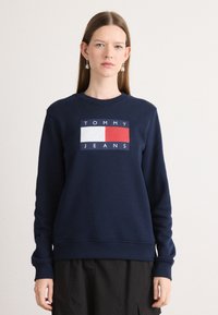 Felpa navy con un logo rettangolare rosso e bianco che riporta la scritta "TOMMY JEANS", polsini a coste e scollatura a girocollo. Tessuto morbido.