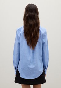 Donna con lunghi capelli castani, che indossa una camicia a righe blu e una gonna nera, in piedi di fronte a un muro grigio chiaro uniforme.