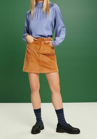 Femme portant un chemisier à manches longues violet clair, une jupe en velours côtelé moutarde, des chaussettes rayées bleu marine et des mocassins noirs, debout devant un fond vert.