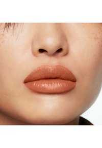 Gros plan sur des lèvres ornées d'un rouge à lèvres nude mat aux teintes chaudes. La peau semble lisse avec une texture naturelle et de légères taches de rousseur.