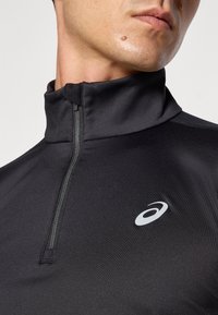 Svart quarter-zip genser laget av glatt, lett stoff. Har en høy krage og en liten, hvit merkevarelogo på brystet.