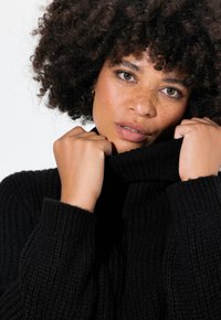 Maglione nero a collo alto lavorato a maglia con texture a costine; il modello tiene il colletto vicino, mostrando la pelle e i capelli ricci su uno sfondo neutro.