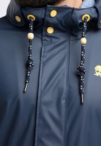 Navy rain jacket with a high collar, featuring yellow snap buttons, two drawstrings with beads, and a logo detail. Smooth, waterproof material.