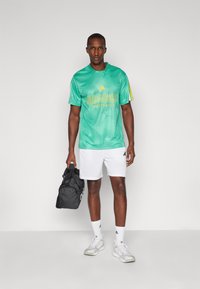 Camisola atlética de malha verde com detalhes amarelos e o texto "ADIDAS SPORTSWEAR", combinada com calções brancos e uma mala de desporto preta.
