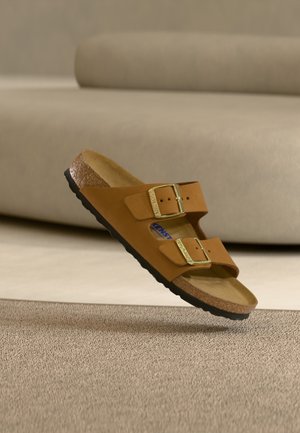 Bruine suède sandaal met twee gouden gespen en kurken zool, zwevend boven een beige tapijt, met een neutrale bank op de achtergrond.