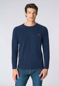 Maglione lavorato a maglia di colore blu navy con maniche lunghe, scollo a girocollo, polsini e orlo a costine, caratterizzato da un piccolo emblema rosso sul petto.