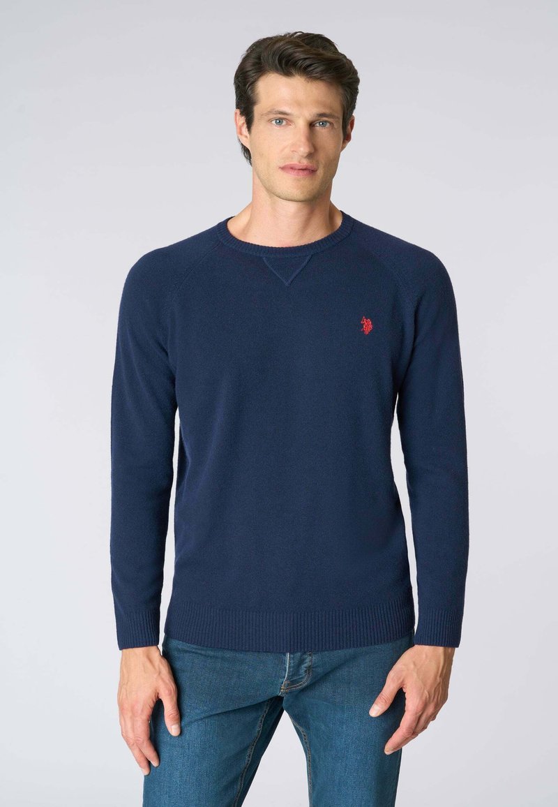 Maglione lavorato a maglia di colore blu navy con maniche lunghe, scollo a girocollo, polsini e orlo a costine, caratterizzato da un piccolo emblema rosso sul petto.