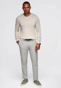 Maglione beige, pantaloni grigi chiari su misura e sneakers grigie. Orologio e braccialetto come accessori. Texture morbide, design aderente.