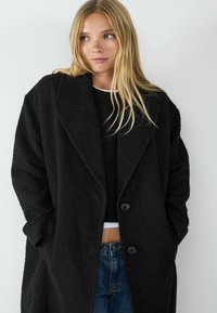 Zwarte oversized jas van een gestructureerde stof, met grote revers en twee knopen. Gedragen over een zwarte cropped top en blauwe jeans.