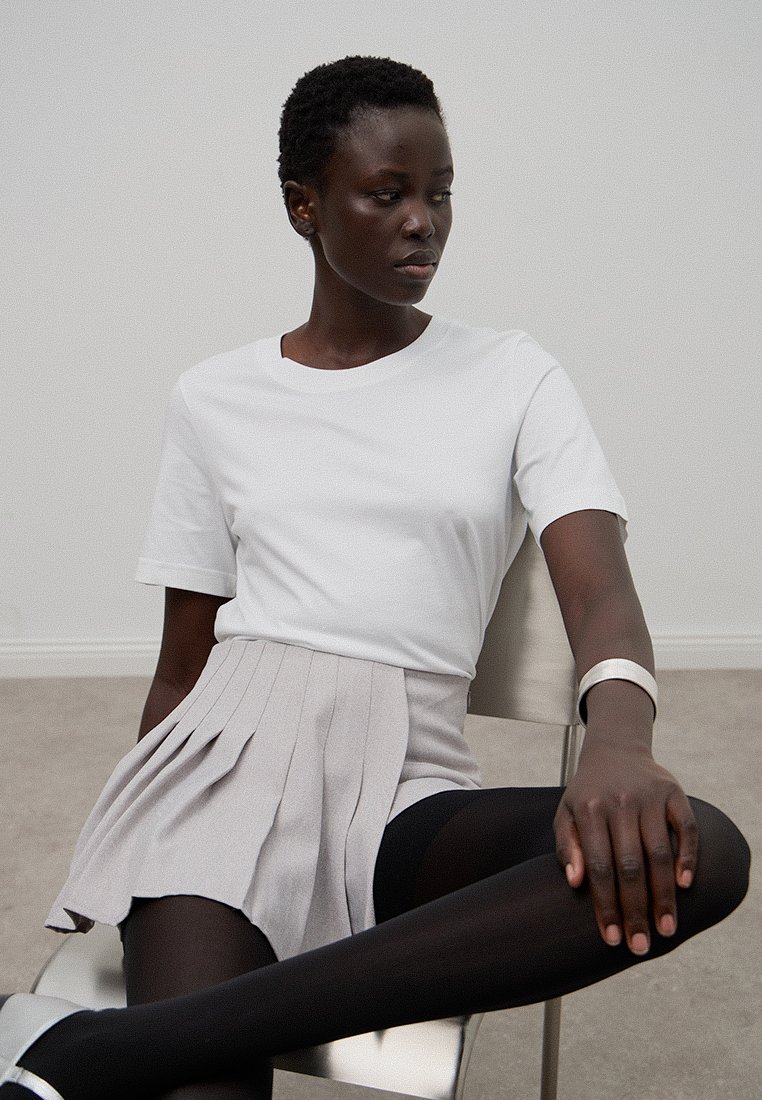 Biała koszulka z krótkim rękawem połączona z jasnoszarym plisowanym mini spódnicą, noszona z czarnymi rajstopami i białą bransoletką na nadgarstku.