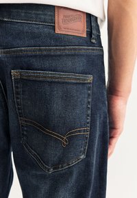 Tasca posteriore di jeans in denim blu scuro con cuciture marroni e una toppa in pelle con la scritta "Premium Denim Goods" vicino alla vita.