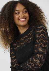 Crna čipkana majica sadrži složene valovite uzorke, prozirne duge rukave i okrugli izrez, ističući teksturirani dizajn i elegantnu izradu.