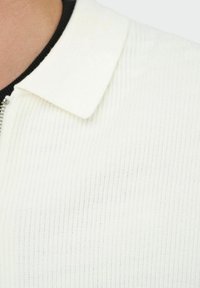 Nahaufnahme eines gerippten weißen Strickpolohemds mit einem Reißverschluss und einem umgeschlagenen Kragen über einem schwarzen Unterhemd bei einer Person.