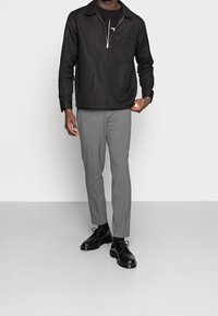 Camisa negra con cremallera y cuello, pantalones gris ajustados, zapatos negros, de pie sobre un suelo gris neutro. Enfoque en los detalles de la tela y el ajuste.