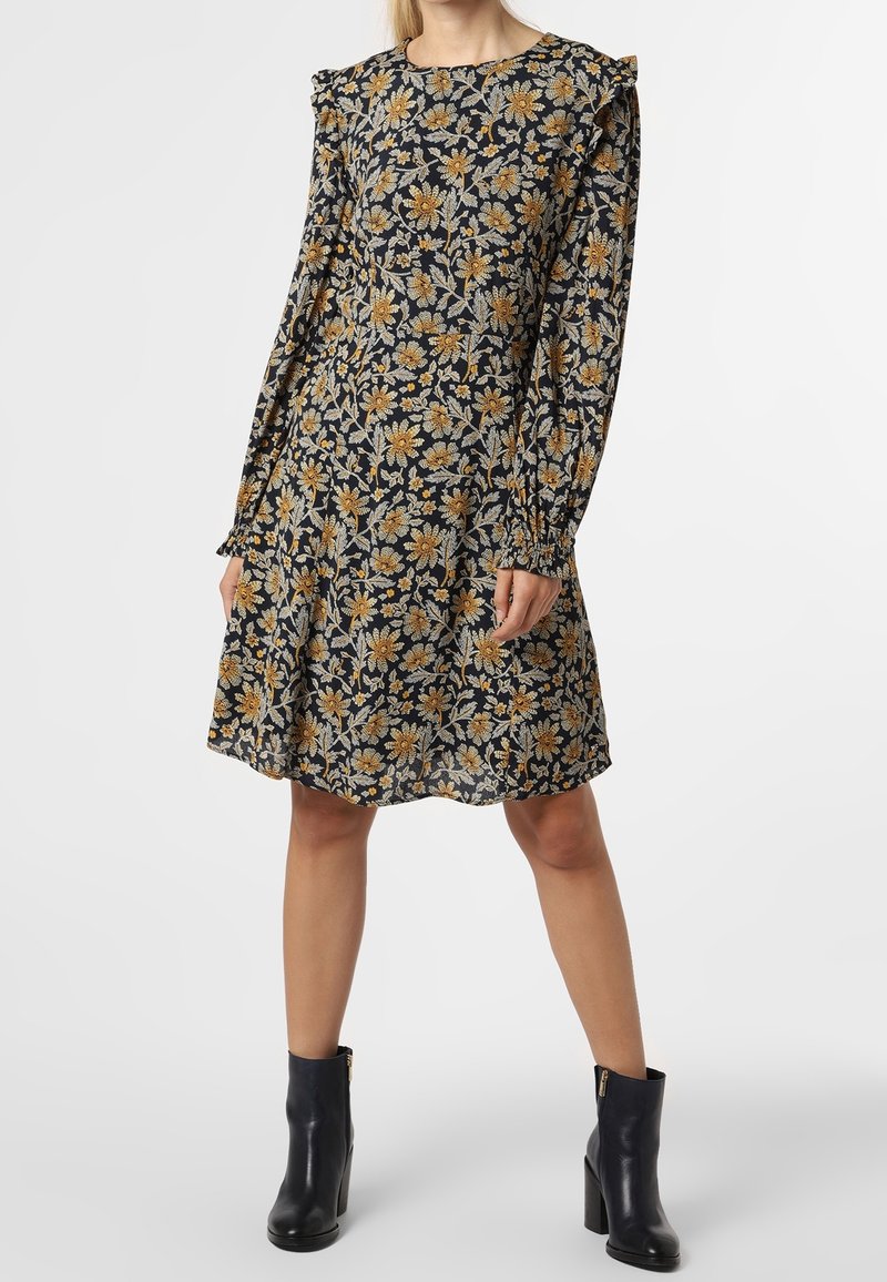 Femme portant une robe noire mi-longue avec un imprimé floral jaune et gris, des manches longues bouffantes et des bottines noires à talons.