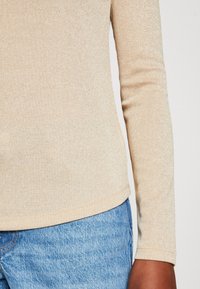 Lange mouw top in beige ribbelstof met een subtiele glans, voorzien van zijsplitten en een afgeronde zoom, gecombineerd met blauwe denim.