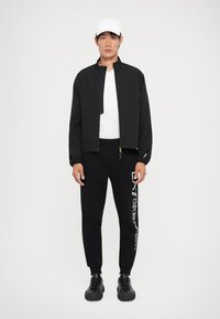Svart jakke med glidelås, hvit T-skjorte med rund hals, svarte joggebukser med hvit logo, og svarte sneakers. Tilbehøret inkluderer en hvit caps.