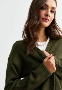 Cardigan avvolgente verde oliva con polsini a costine e scollatura a V profonda, realizzato in materiale morbido e testurizzato, abbinato a una camicia bianca a strati.