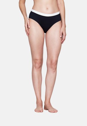 Kobieta w czarnej bieliźnie z białym paskiem, stojąca boso na jasnym, jednolitym tle, widoczna od talii do stóp.