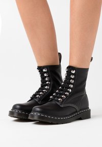 Botas de tobillo de cuero negro con ojales plateados, diseño de cordones, detalles de costura y una suela de goma gruesa. Aptas para diversos estilos.