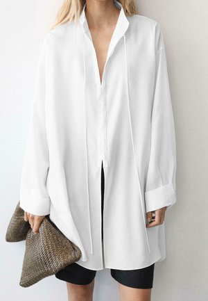 Femme portant une chemise blanche ample à manches longues tenant une grande pochette tissée marron, associée à un short noir.