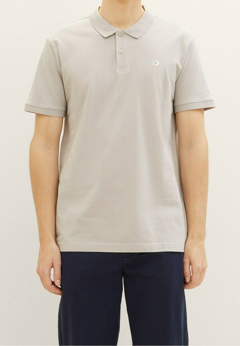 Homme portant un polo gris clair à manches courtes et un pantalon bleu marine foncé, debout devant un fond clair uni.