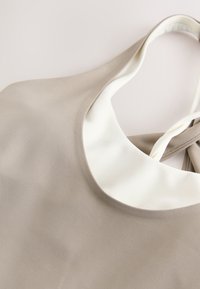 Sleeveless beige top with a white round neckline and a thin strap tied at the back.