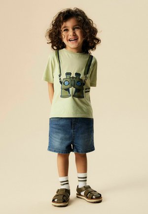 Unge barn med krøllet hår smiler, iført lysegrøn t-shirt med kikkertprint, denimshorts, stribede sokker og brune sandaler, stående.