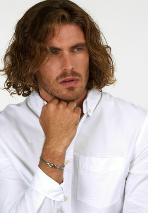 Camicia bianca con bottoni, dotata di taschino sul petto, caratterizzata da un braccialetto in catena d'argento al polso, capelli ricci castani e uno sfondo neutro.