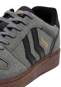 Szare zamszowe sneakersy z czarnymi akcentami, z trzema paskami, szarymi sznurowadłami i teksturowaną brązową gumową podeszwą. Złote logo marki po boku.