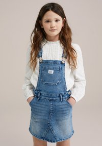 Denim overal obleka z modrim pranjem, z nastavljivimi naramnicami, sprednjimi žepi in raztrganim robom. V kombinaciji z belo strukturirano majico z dolgimi rokavi.