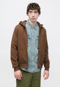 Καφέ φούτερ με φερμουάρ και τσέπη καγκουρό, φορεμένο πάνω από ένα ανοιχτό μπλε πουκάμισο από τζιν. Διαθέτει κουκούλα με κορδόνι και ελαστικές μανσέτες.