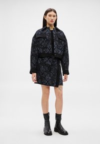 Femme portant une veste texturée noire et bleu foncé et une jupe assortie, avec des bottines à talons noires, debout sur un fond blanc.