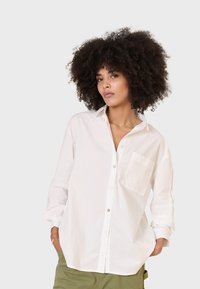 Camicia bianca oversize in cotone con taschino, dettagliata con bottoni verdi e maniche arrotolate. Texture liscia.