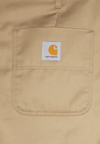 Beige tkanina s texturovaným, diagonálním tkaním obsahuje zadní náprsní kapsu. Čtvercová bílá nálepka zobrazuje žluté logo a text "carhartt".
