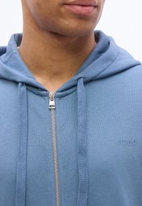 Κοντινό πλάνο ενός ατόμου που φοράει ανοιχτό μπλε φούτερ με φερμουάρ και κορδόνια, με μικρό λογότυπο "HUGO" στο στήθος.