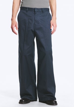 Mann trägt dunkle, weite Hosen und schwarze Schuhe, steht vor einem schlichten hellen Hintergrund.