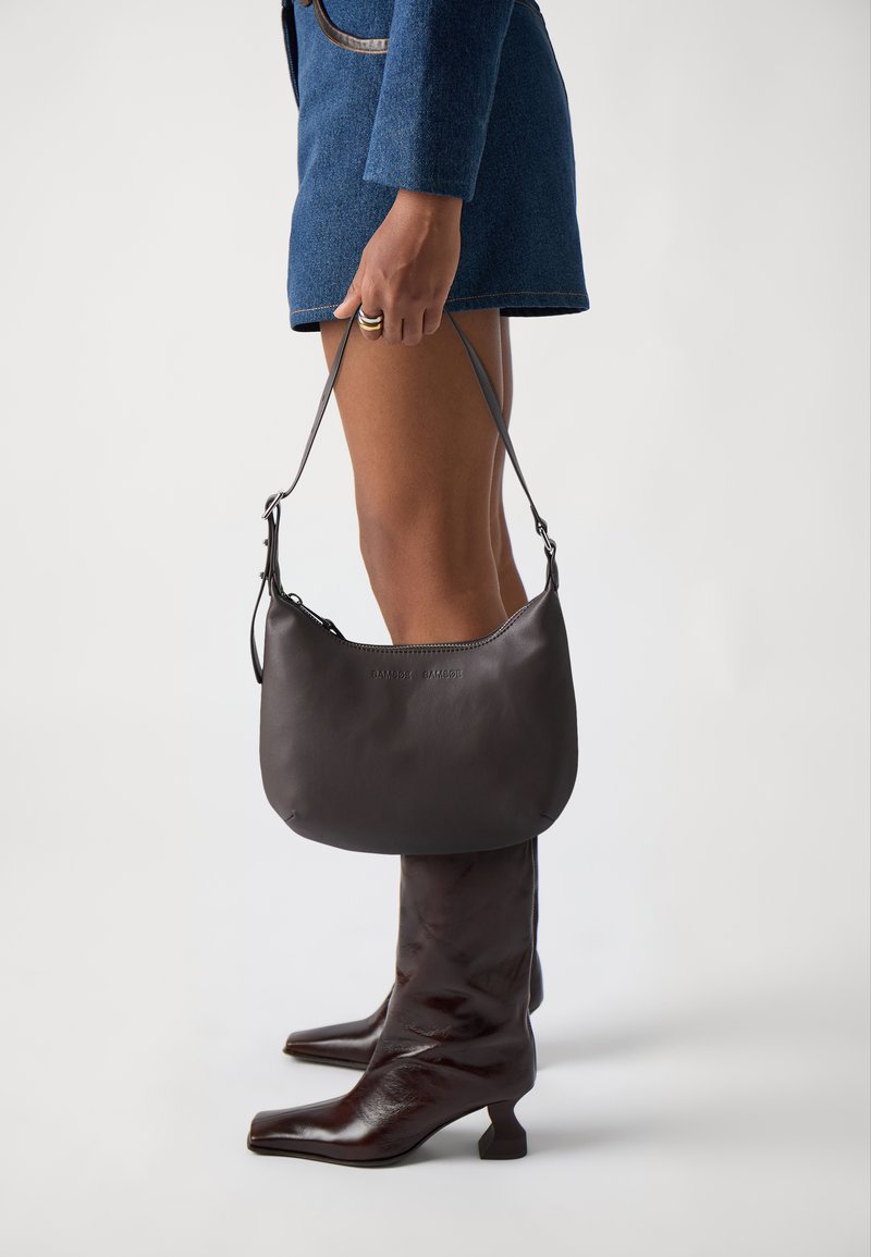 Porte-monnaie en cuir noir avec une forme incurvée et une bandoulière ajustable, tenu par une personne portant des bottes marron jusqu'aux genoux, sur un fond neutre.