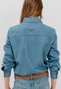 Jasnoniebieska koszula dżinsowa, z długimi rękawami i zapięciem na guziki, z detalem szwu w karczku, minimalistyczny design, noszona z brązowym skórzanym paskiem.
