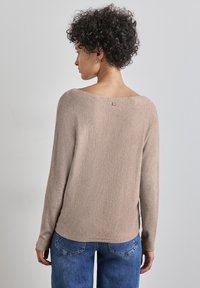 Beige langärmliger Pullover mit Rundhalsausschnitt und strukturiertem Finish, versehen mit einem kleinen Logopatch auf der Rückseite, kombiniert mit blauen Jeans.