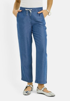 Mujer con pantalones azules de cordón, zapatos planos Mary Jane plateados y una blusa blanca sin mangas con las manos en los bolsillos.