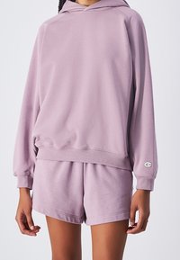 Felpa viola chiaro con cappuccio regolabile, polsini e orlo a coste, abbinata a pantaloni corti coordinati. Tessuto morbido e testurizzato. Logo Champion sulla manica.
