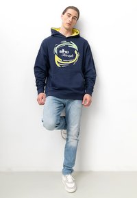 Marineblauer Hoodie mit gelbem Innenfutter und kreisförmigem Grafikelement, kombiniert mit hellblauen Jeans und weißen Sneakers. Glatter Stoff, lässige Passform.